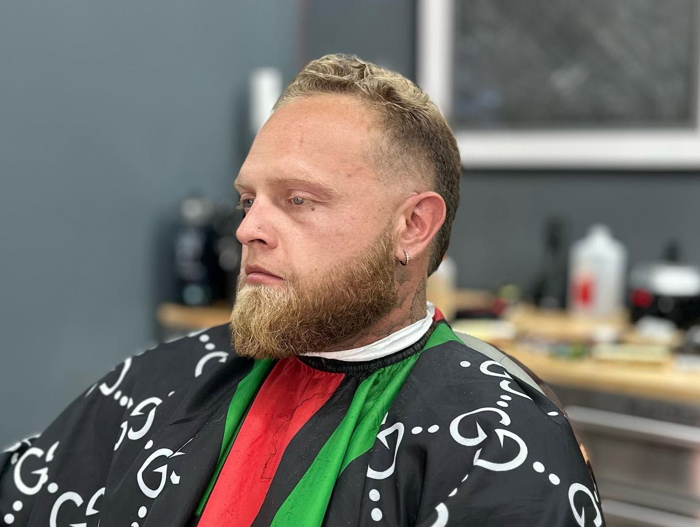 Man with beard in a red-and-green barber cape, seated in a salon chair looking left.