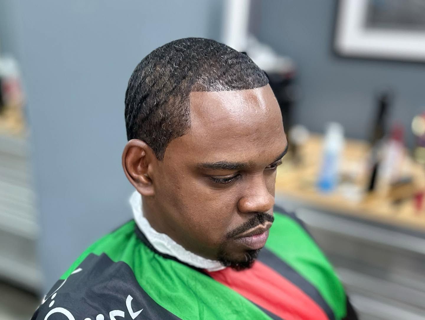 Man in barber chair wearing a green, black, and red cape, looking down in a barbershop