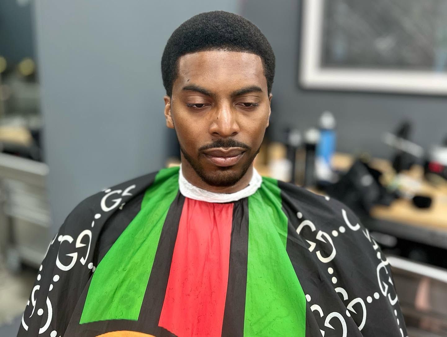 Person seated indoors wearing a black, red, and green barber cape, looking down thoughtfully