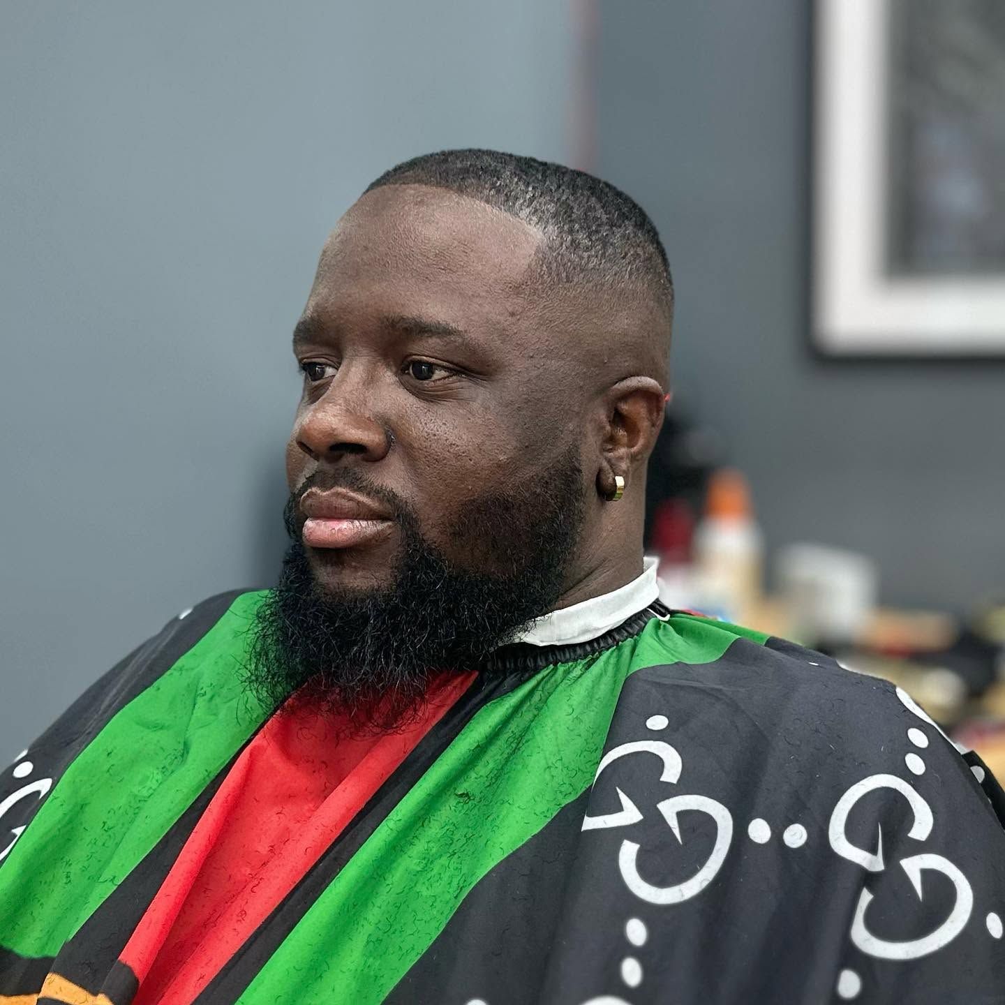 Man seated in a barber chair wearing a green and red cape, with a trimmed beard and short haircut.