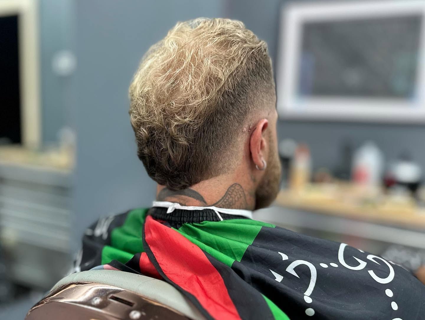Person with blond hair sitting in a chair, seen from behind, wearing a black-and-red jacket in a room
