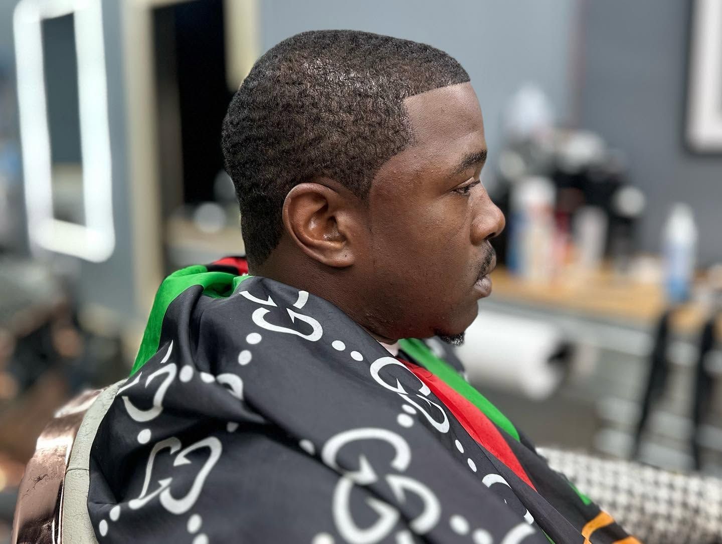 Man seated in a barber chair wearing a black cape with white logos, getting a haircut in a barbershop