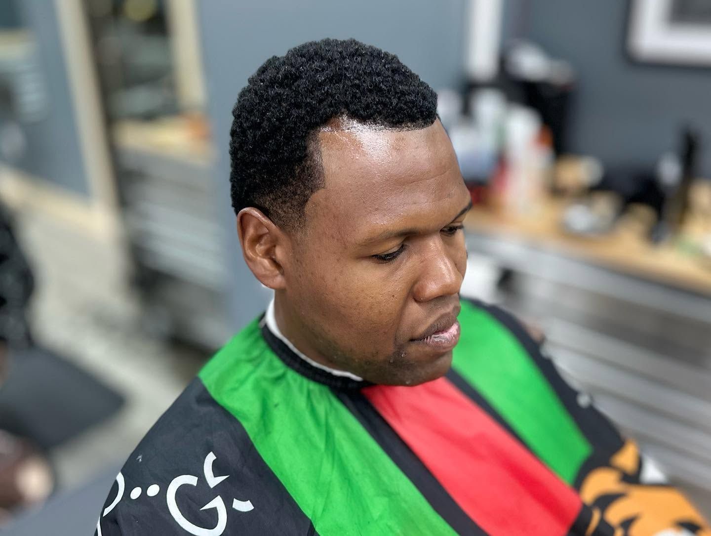 Person seated in a barbershop, wearing a green, red, and black cape, looking down.