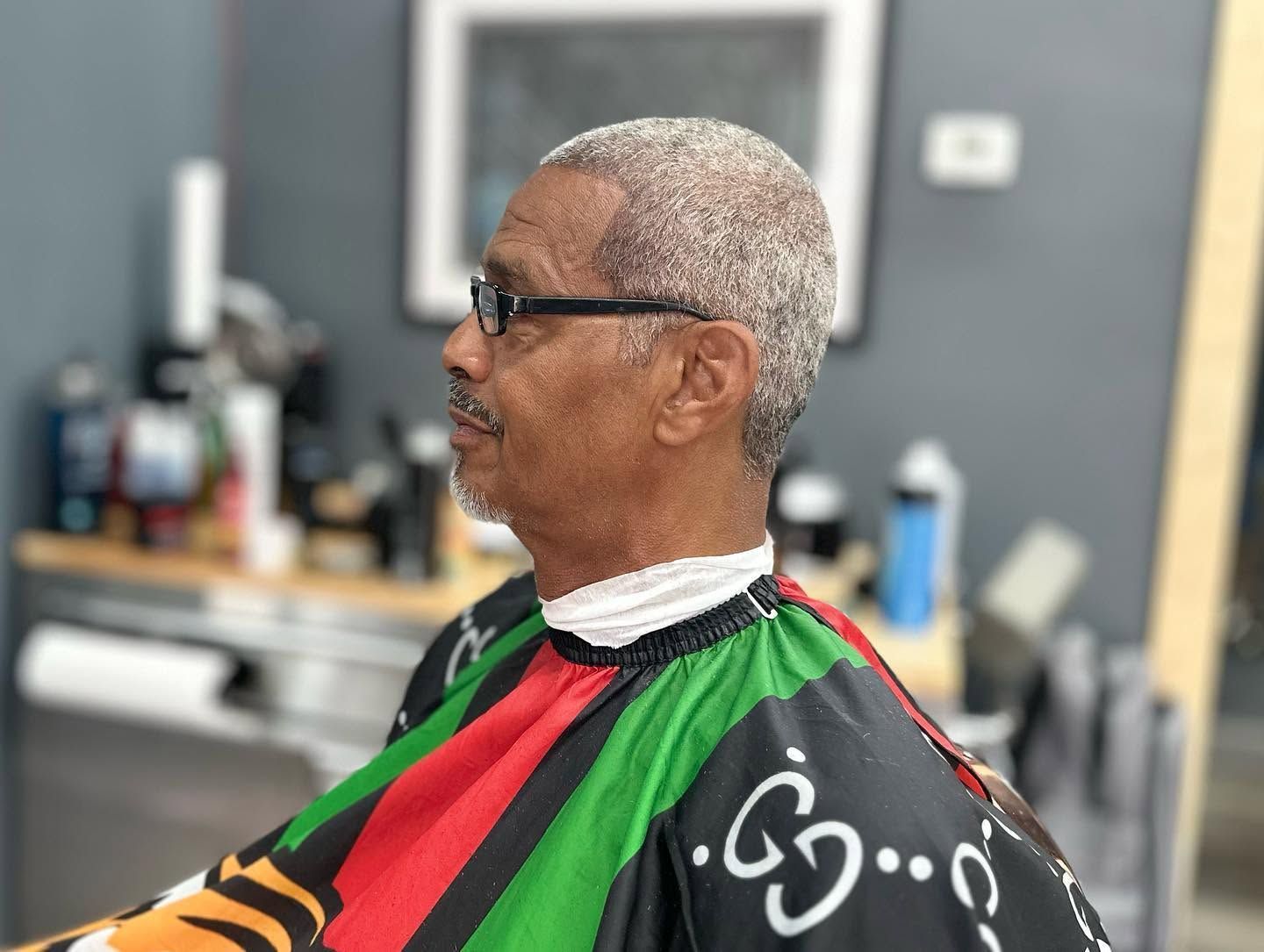 Man in a barber cape seated in profile at a barbershop chair, wearing glasses and short gray hair.