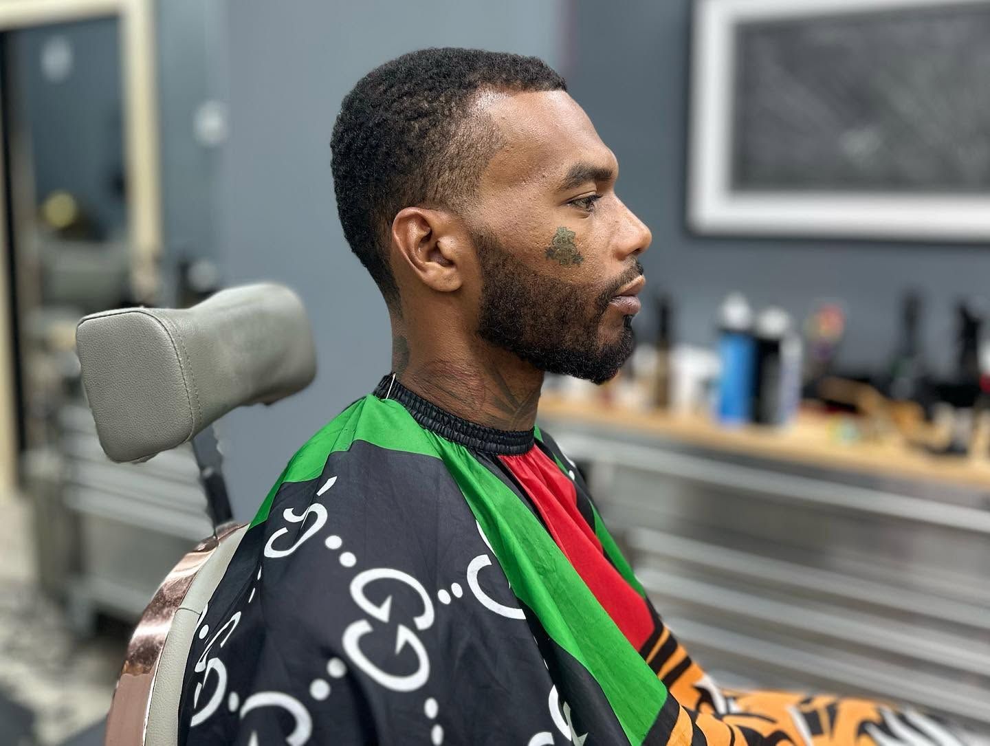 Man seated in a barber chair with a patterned cape in a modern barbershop, shown in profile.