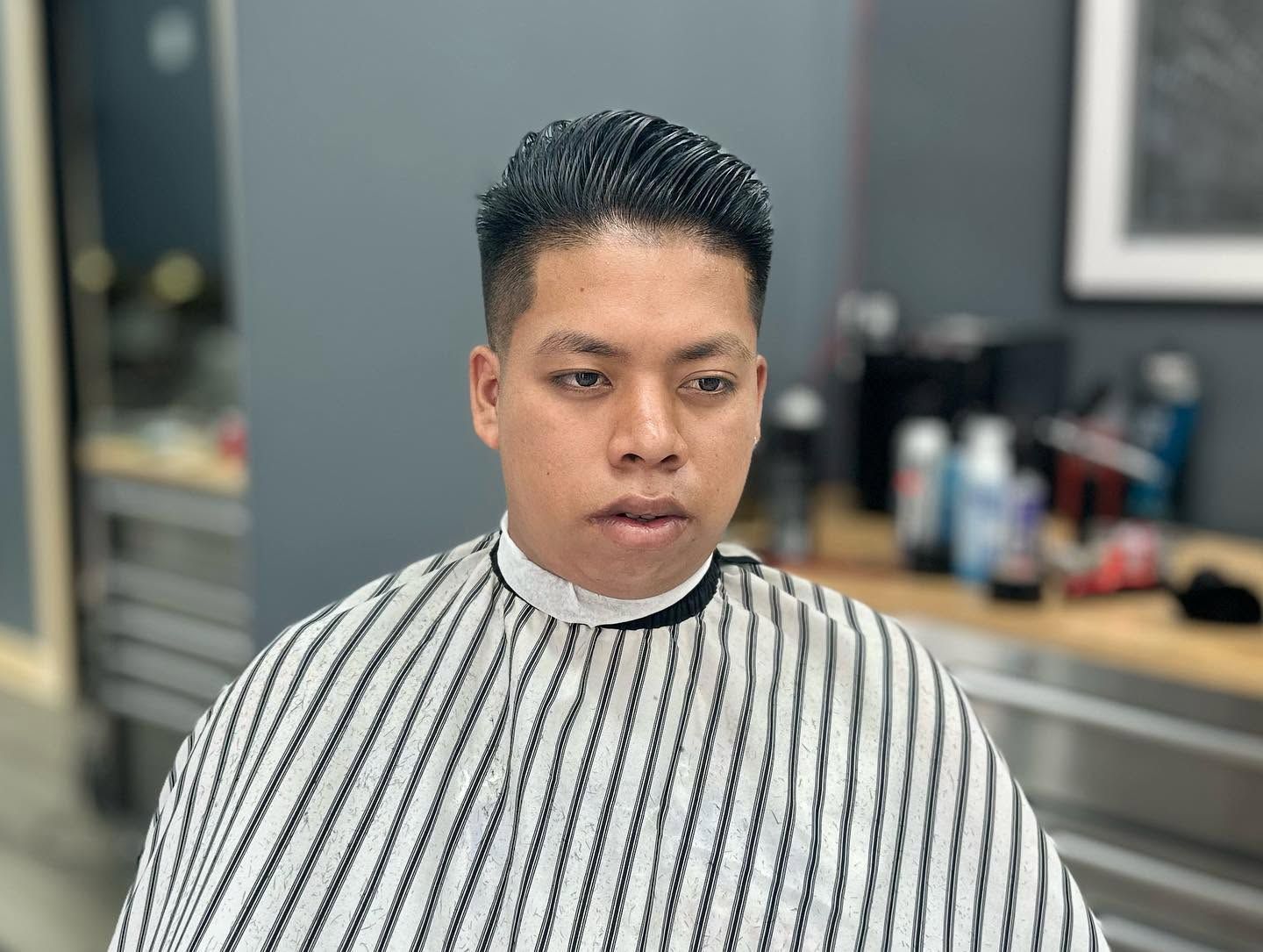 Person with short dark hair wearing a striped barber cape, seated in a salon chair.