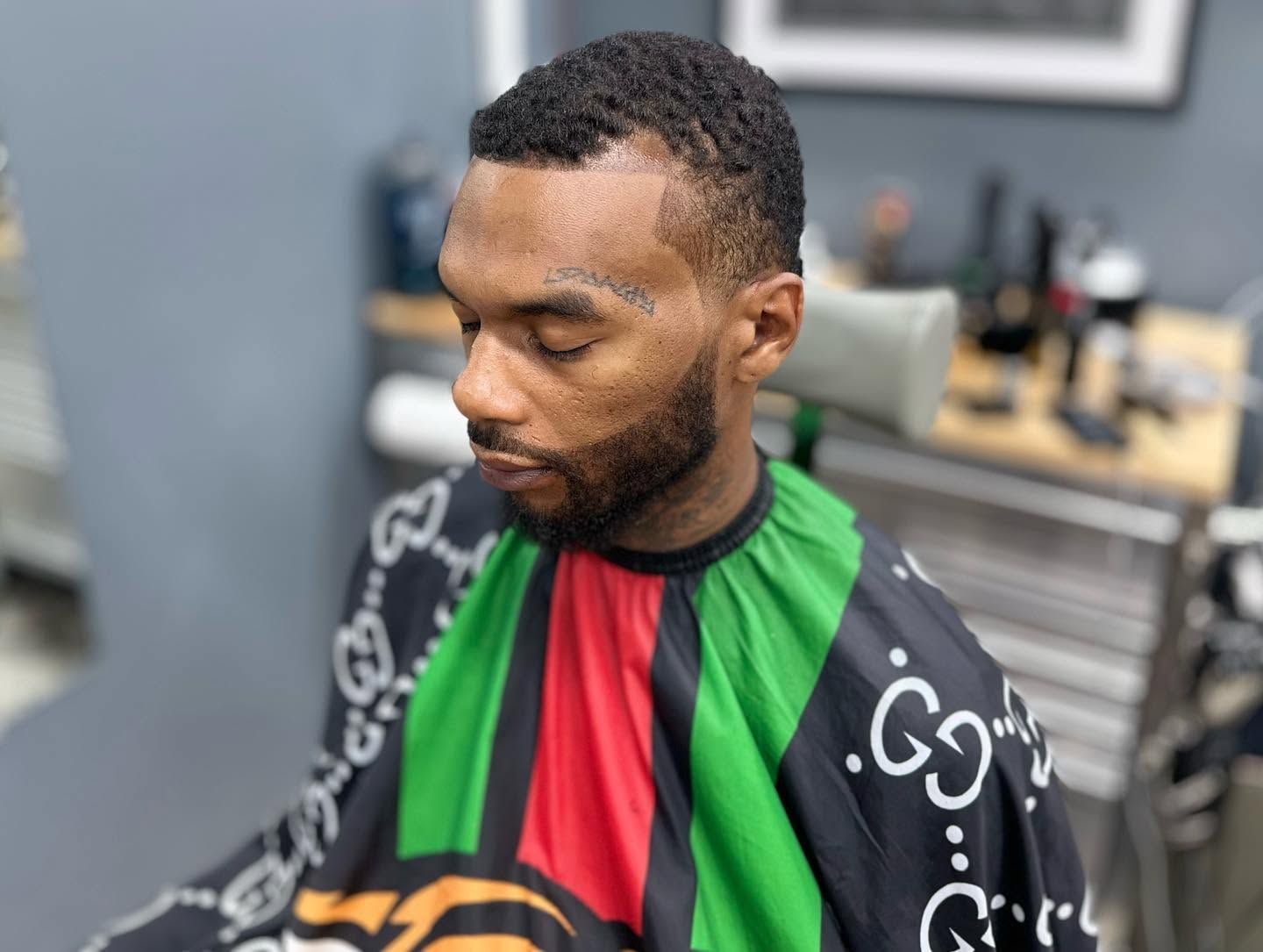 Man in a green, red, and black striped cape sitting in a barber chair, looking down