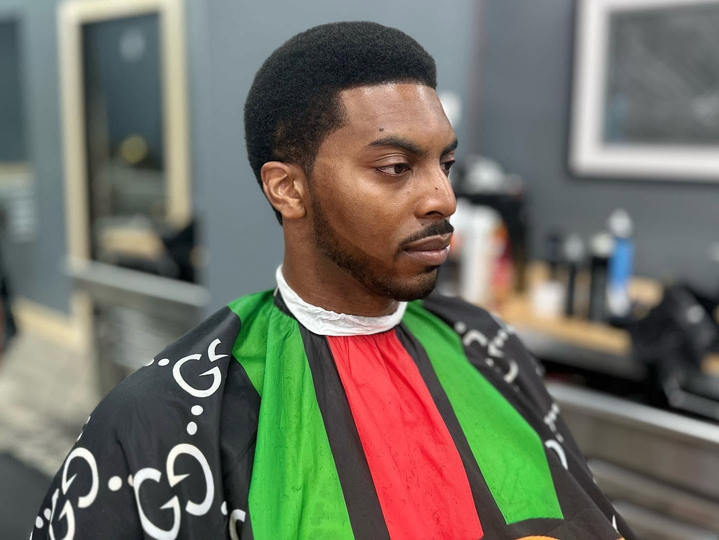 Person in a barber chair wearing a black cape with green and red panels, looking to the side.