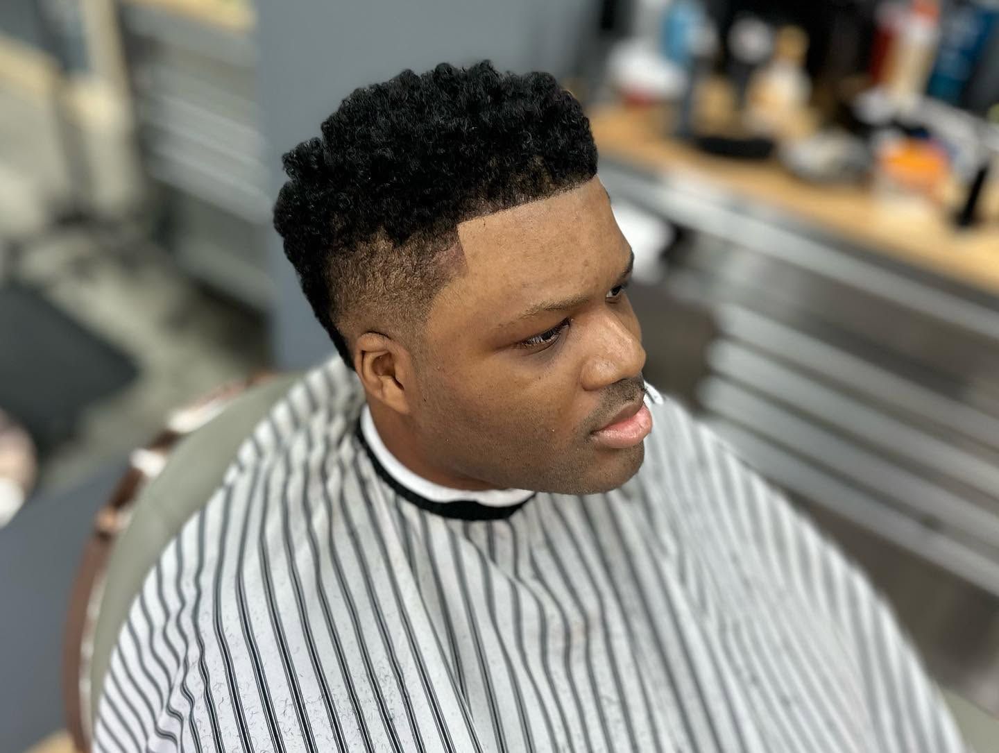 Person with a short curly fade haircut in a striped barber cape, sitting in a barbershop chair and looking to the side