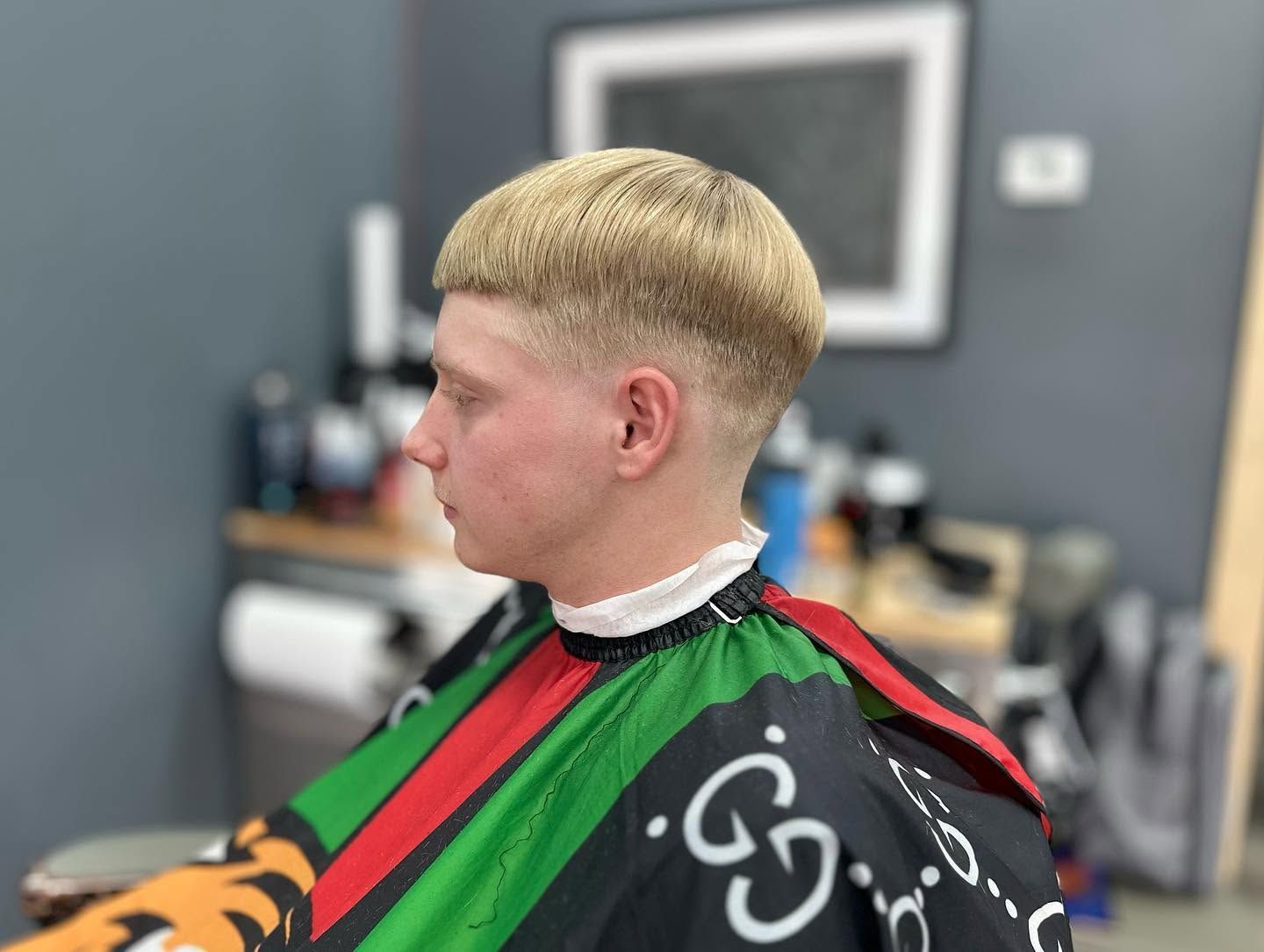 Person with a blond bowl haircut getting a green and red barber cape in a barbershop