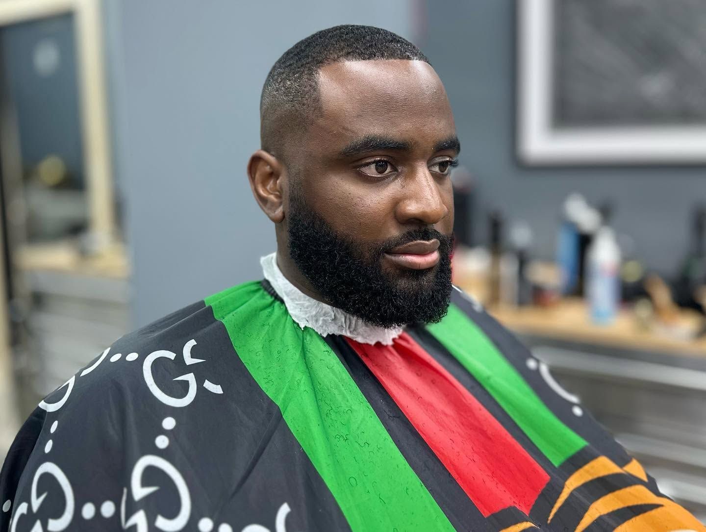 Man seated in a barber chair wearing a black cape with green, red, and yellow stripes in a barbershop