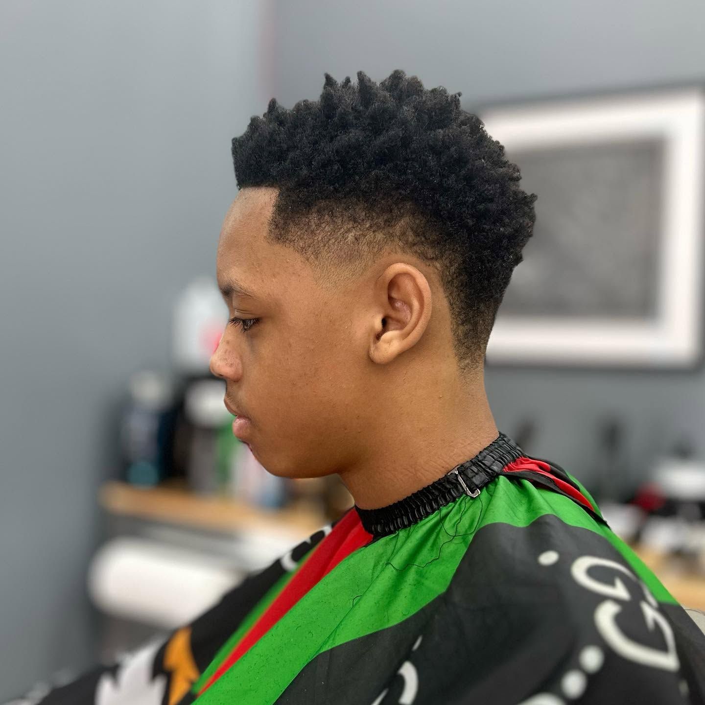 Profile view of a person with a short curly haircut wearing a green and black barber cape in a salon