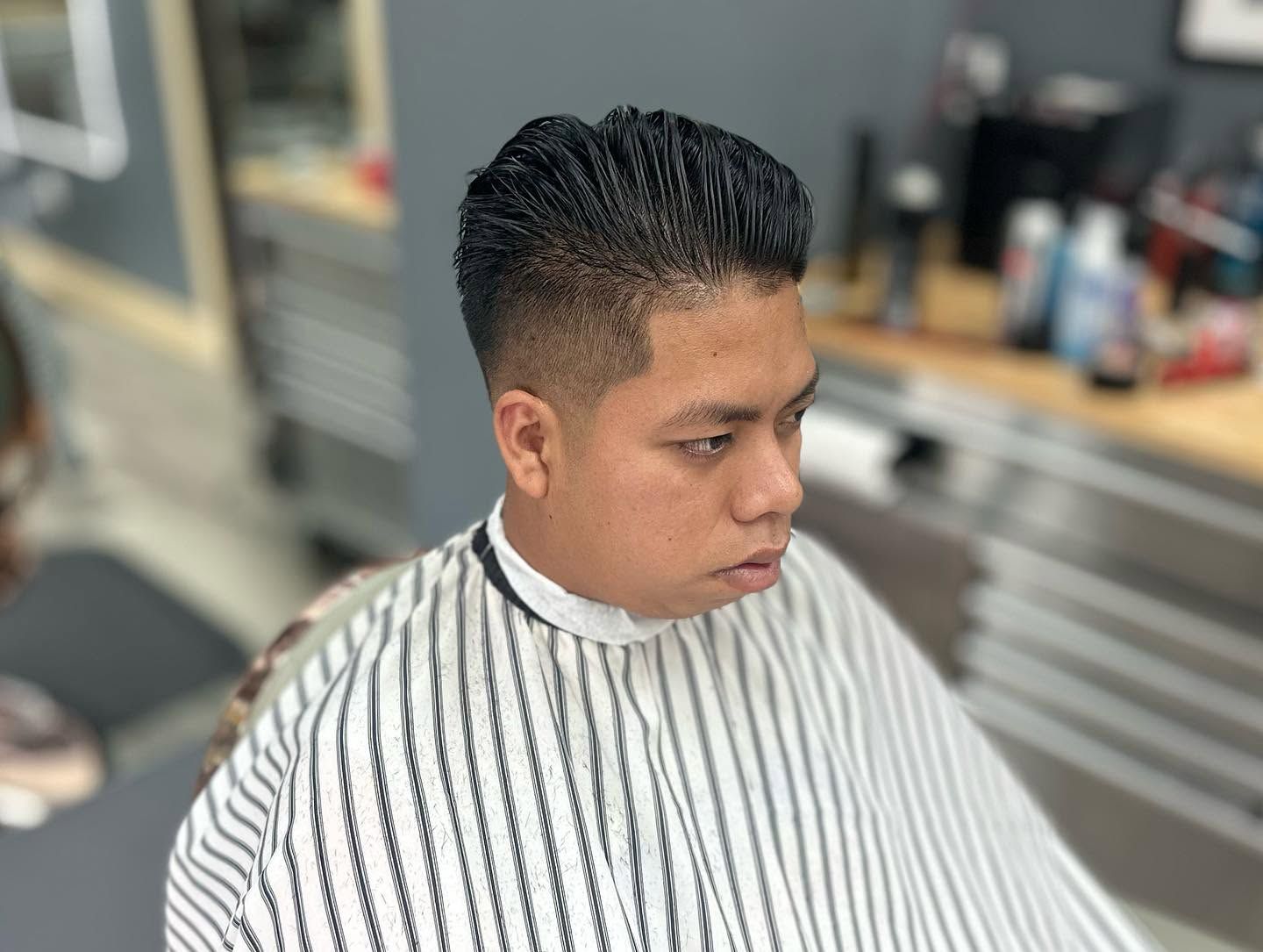 Person in a barbershop cape with slicked-back hair, looking to the side during a haircut.