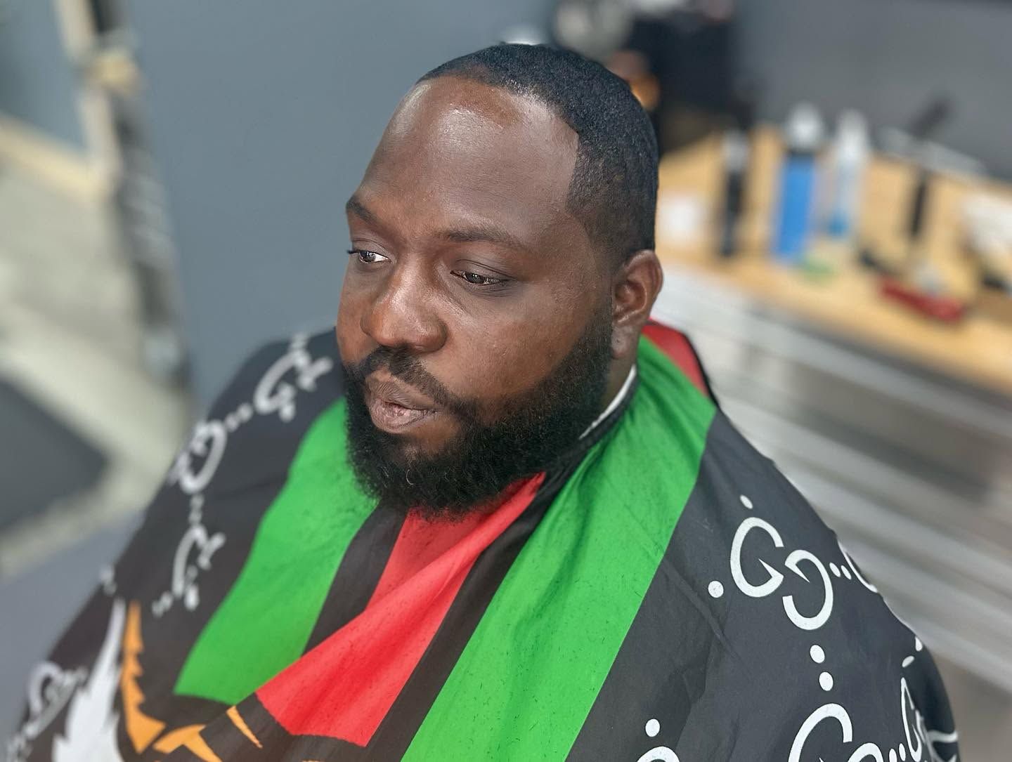Man seated in a black barber cape with green and red stripes, looking down in a barbershop