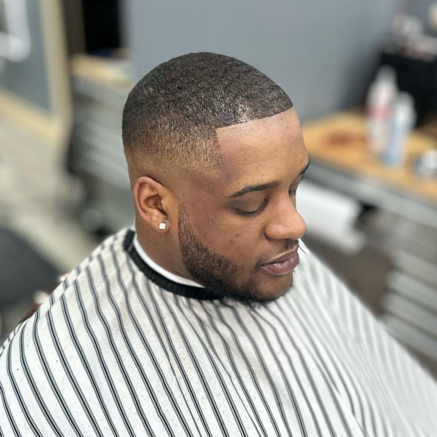 Man with a close-cropped haircut and beard in a black-and-white striped barber cape, looking down in a barbershop.