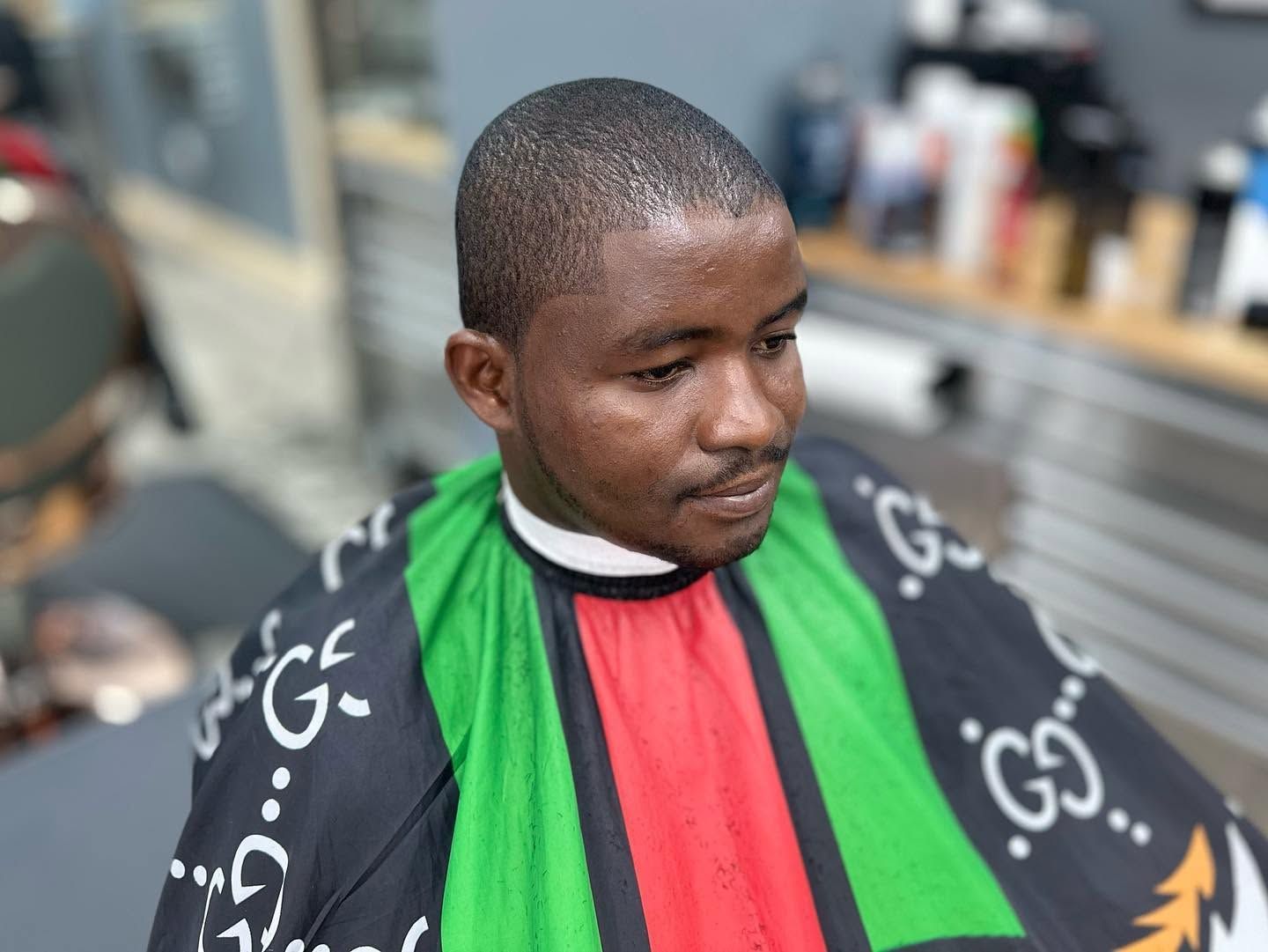Person seated in a barbershop, wearing a green, red, and black cape, looking down.