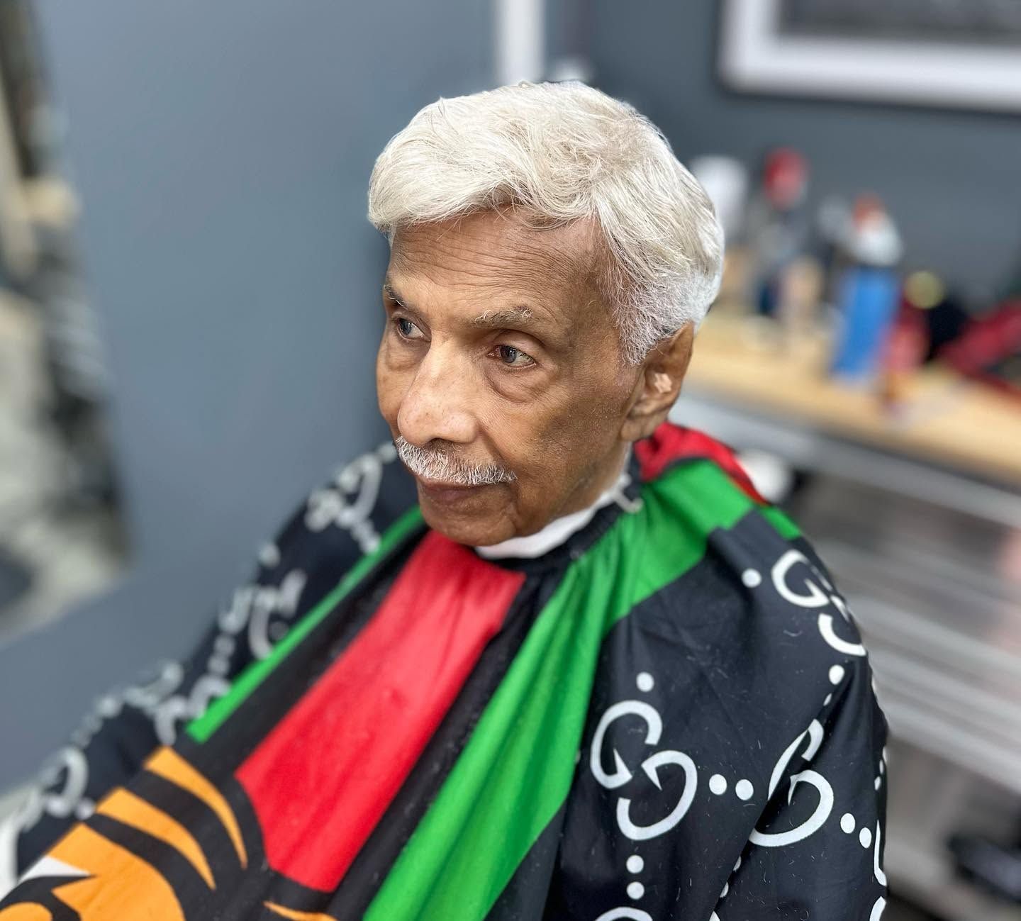 Elderly man with white hair and mustache wearing a black, red, and green barber cape in a salon