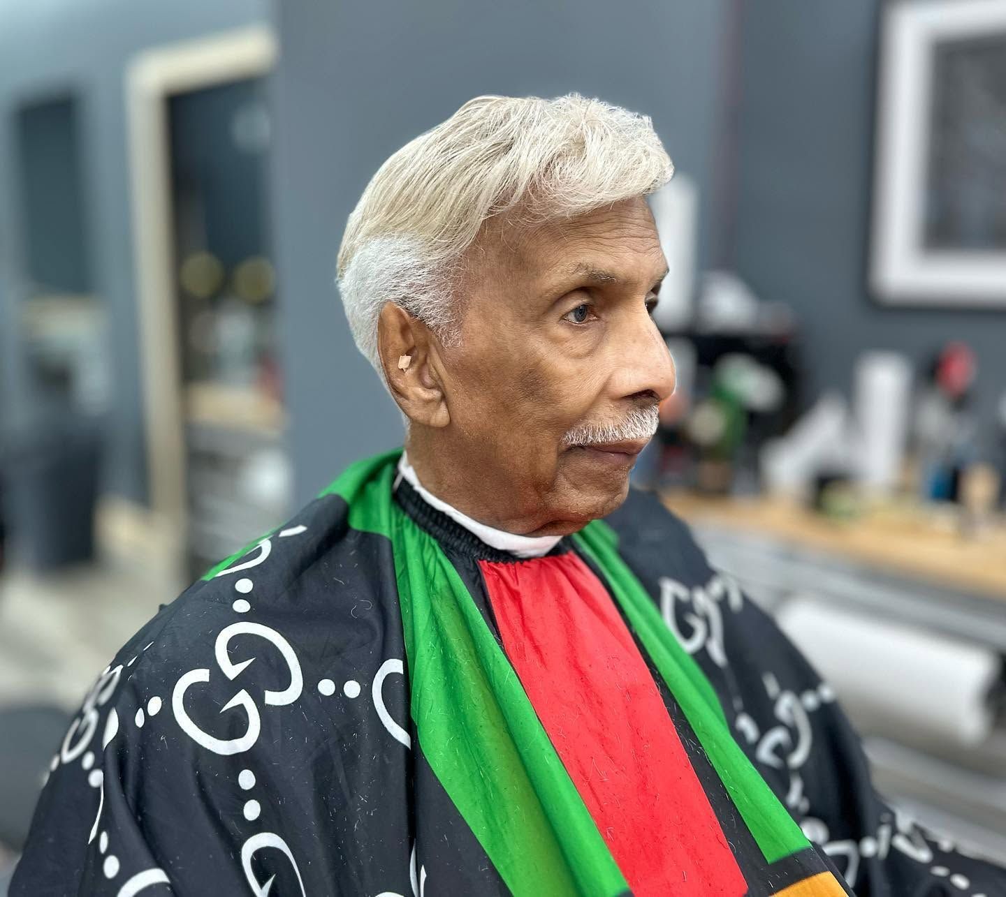 Older man in a black patterned shawl with red and green stripes, seated indoors and looking to the right