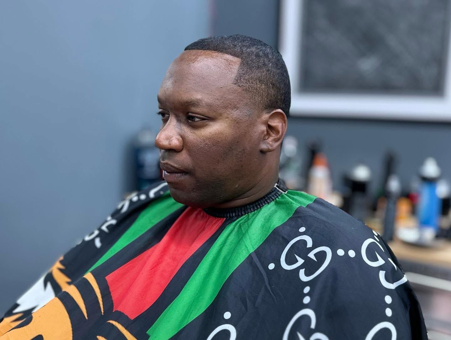 Man wearing a black, red, and green barber cape, seated in a salon chair, looking to the side.