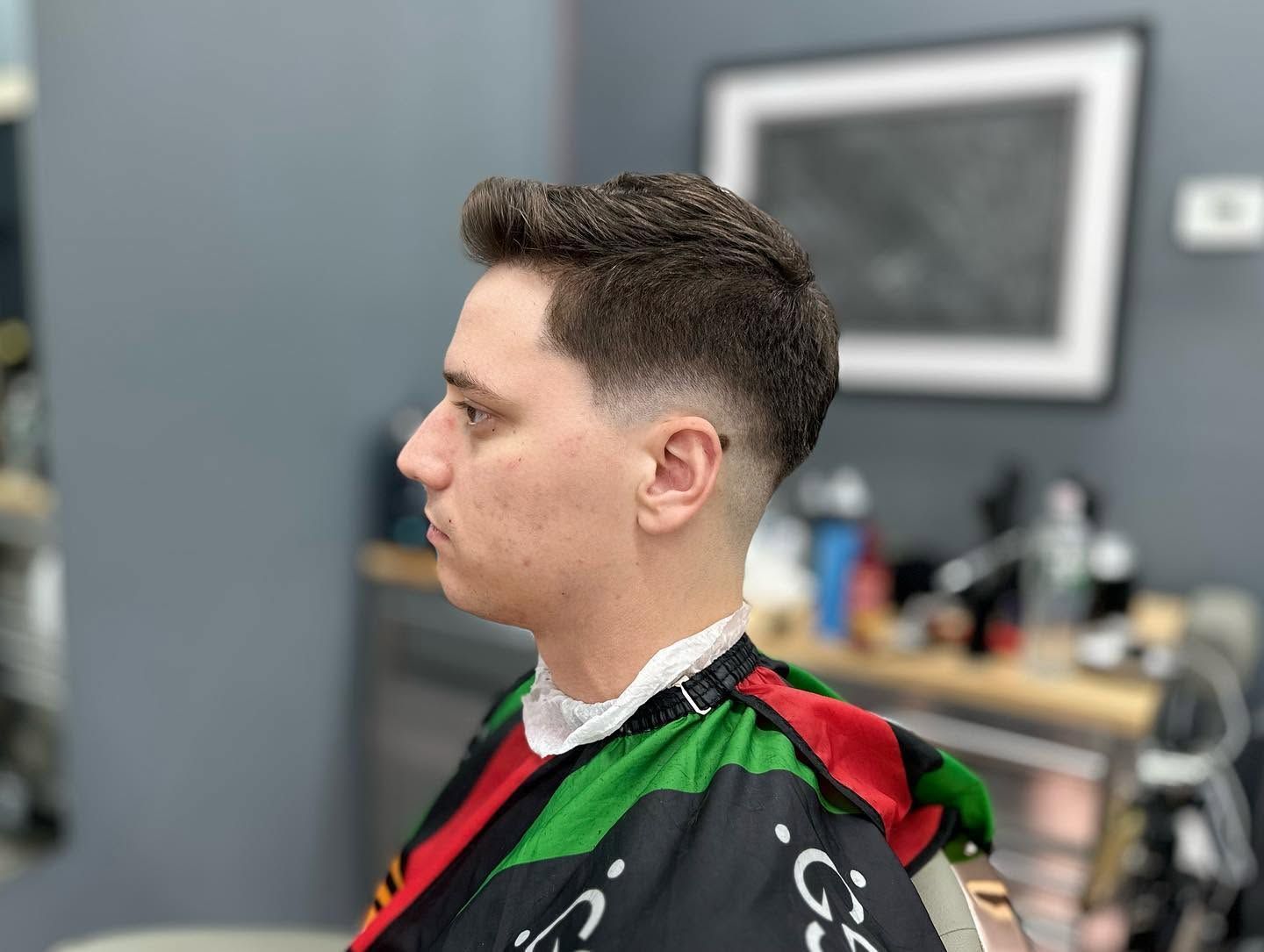 Side profile of a person in a barber chair wearing a black cape with red and green trim in a salon.
