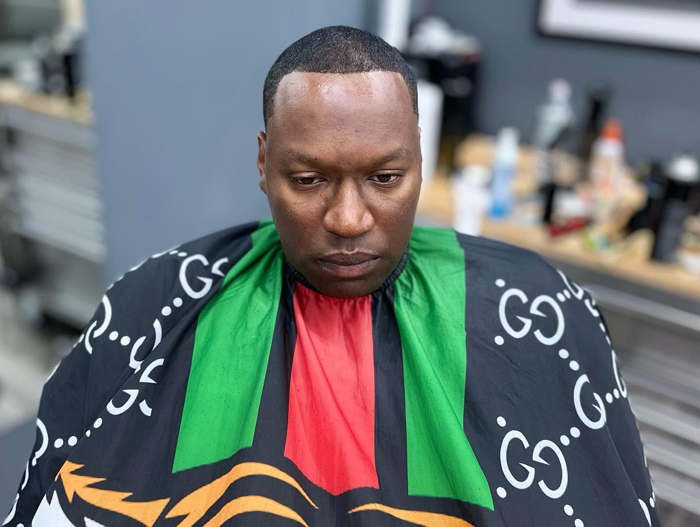 Person seated in a barber chair wearing a black cape with green and red stripes, looking down