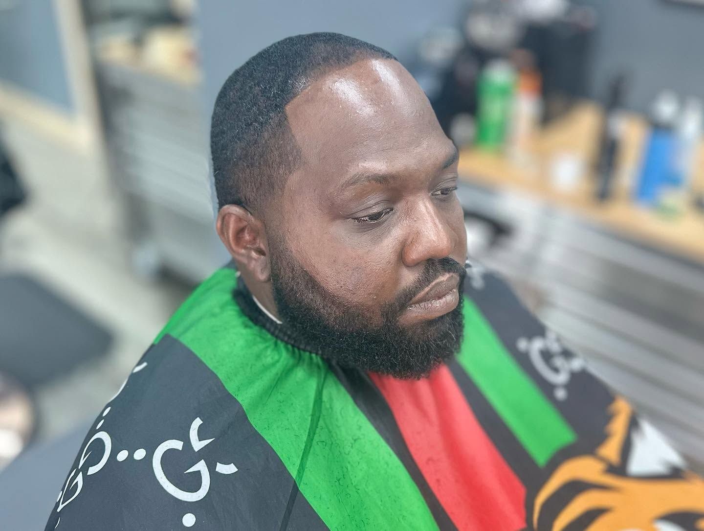 Man in a green, black, and red barber cape, seated and looking to the side in a salon