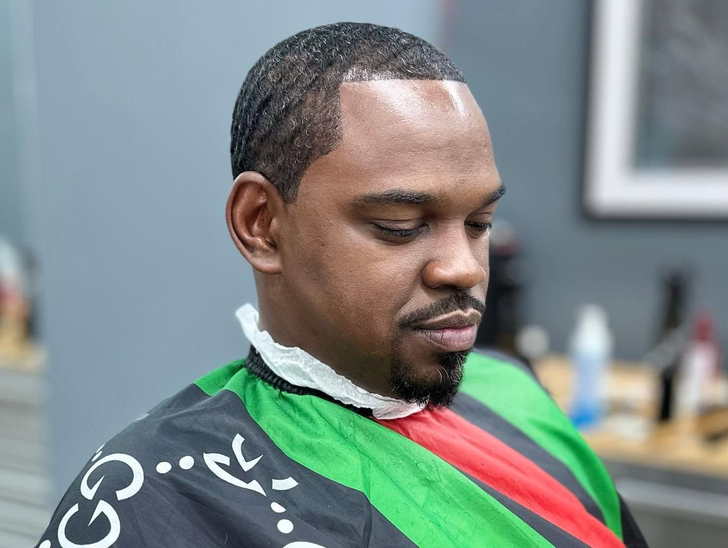 Man getting a haircut in a barber chair, wearing a green and red cape.