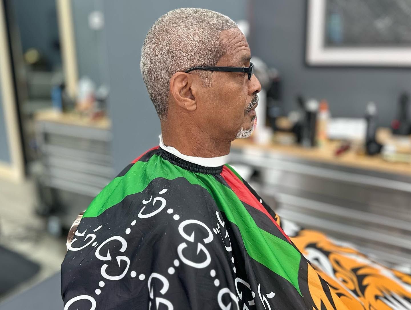 Man seated in profile at a barbershop, wearing a black cape with white patterns and green-red trim.