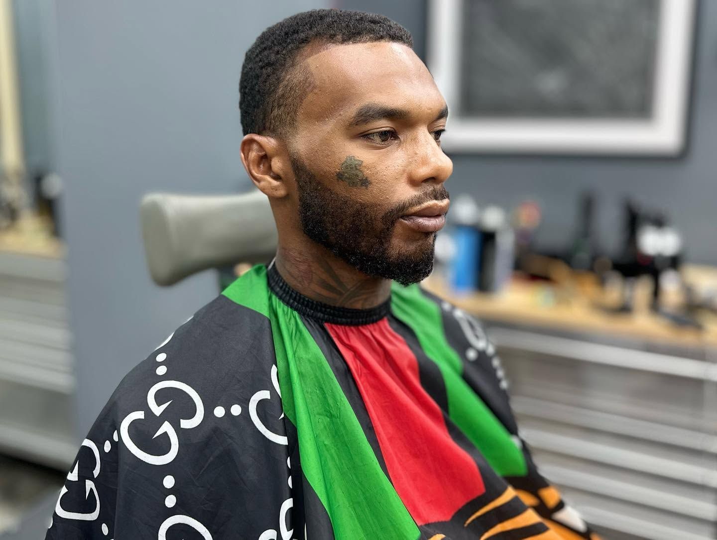 Man seated in a barber chair wearing a red, green, and black cape in a salon or barbershop.