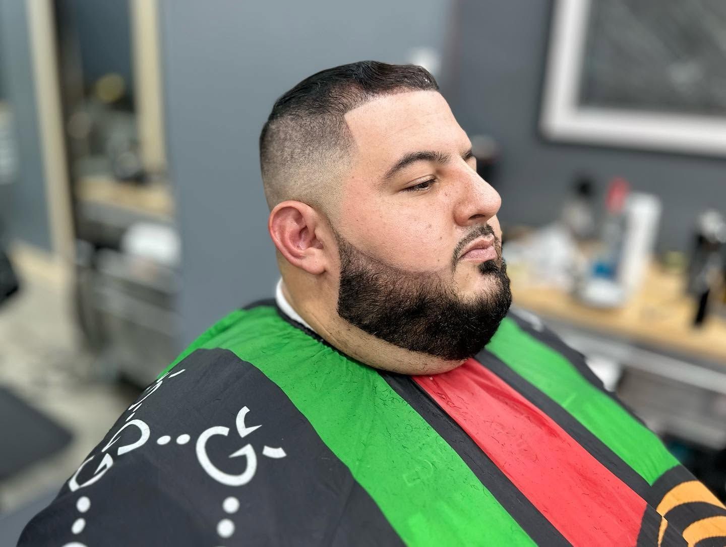 Man seated in a barber chair wearing a black cape with green and red stripes, eyes closed in a barbershop.