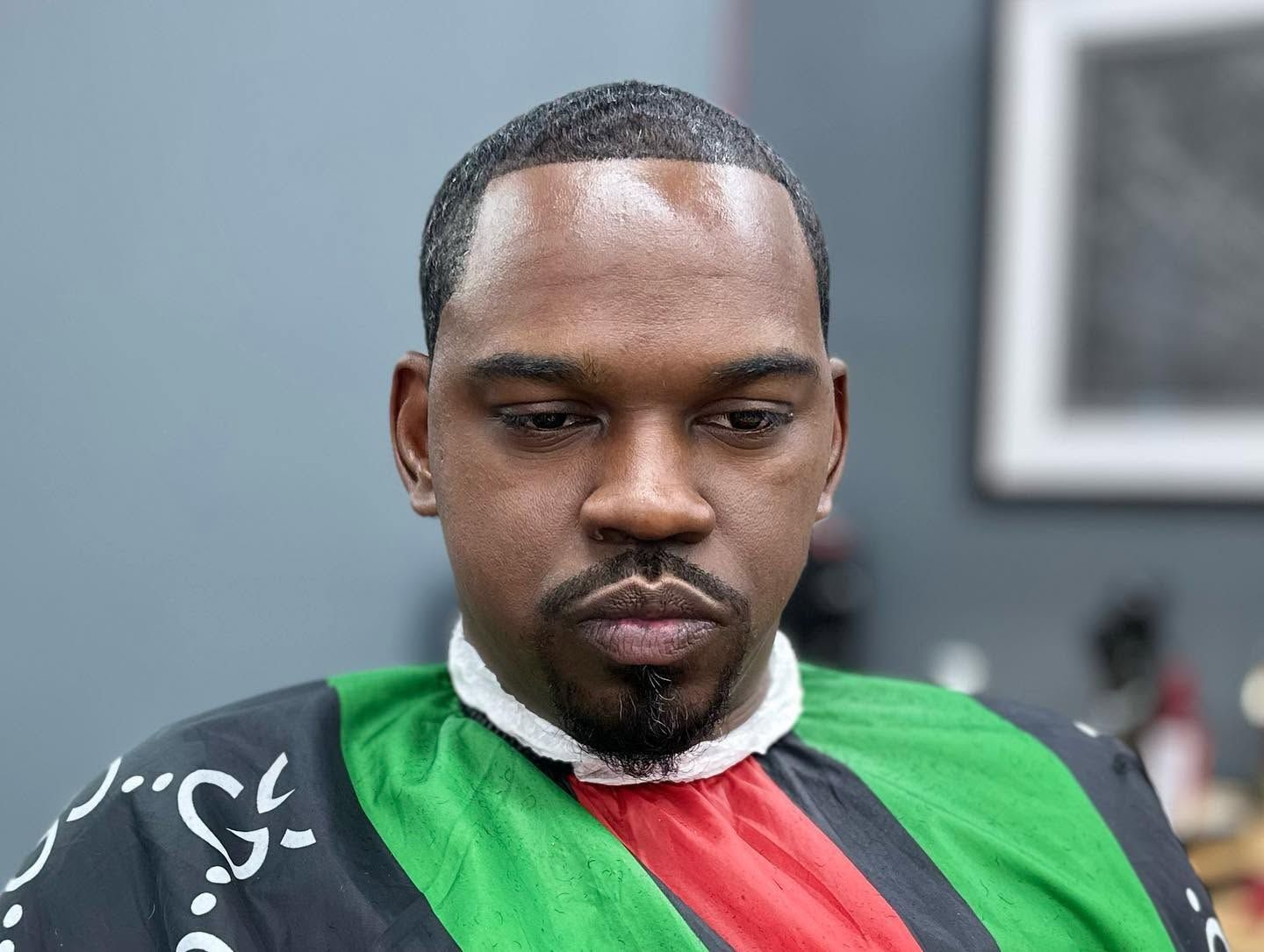 Portrait of a man in a green, red, and black barber cape, looking down in a salon chair