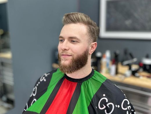 Man with beard in a green and red barber cape, seated in a barbershop chair