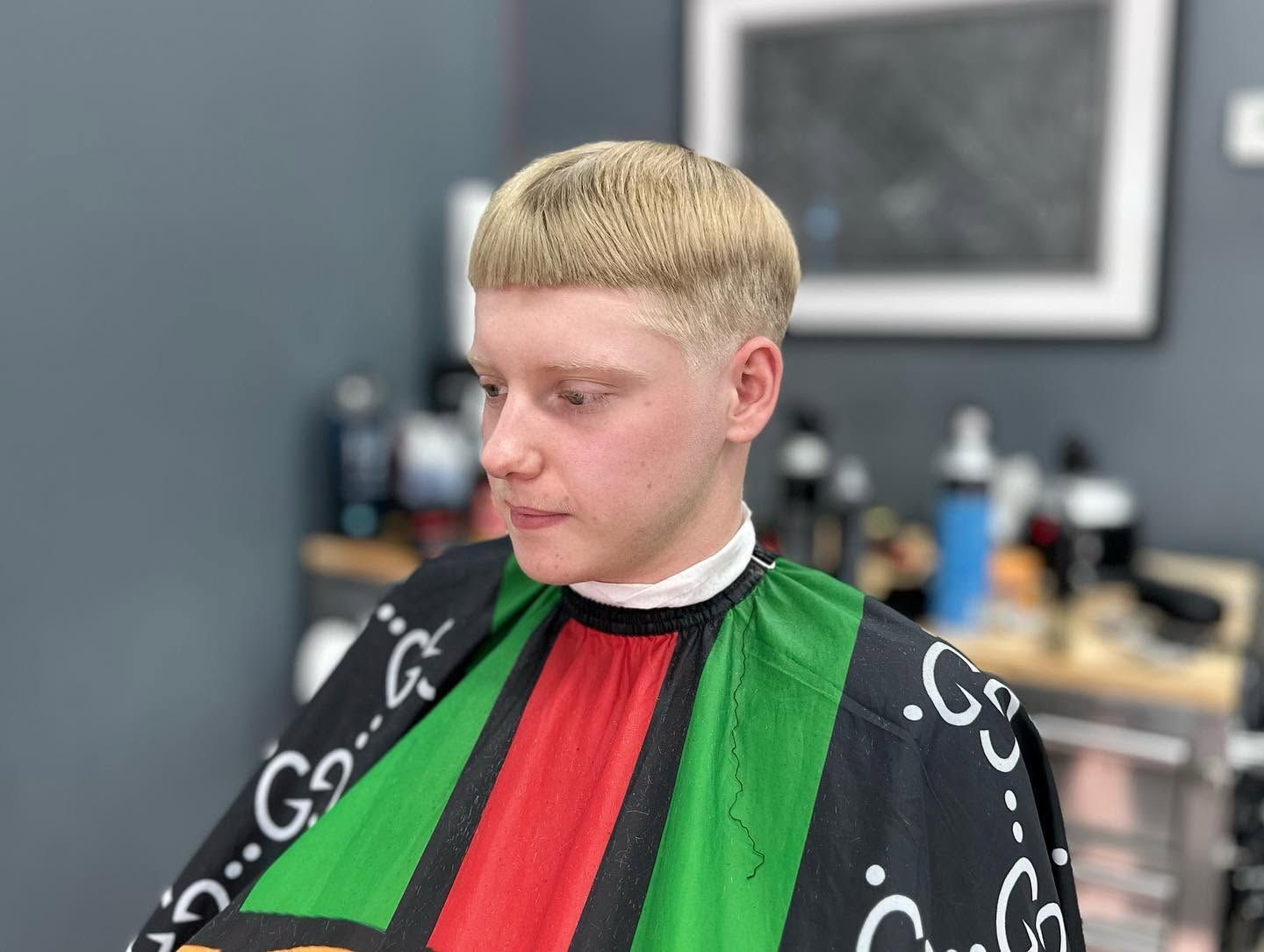 Person in a barber chair wearing a black, green, and red cape in a salon, with short blonde hair.