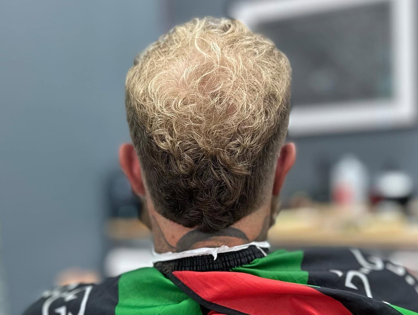 Back of person with short curly blond hair, wearing a black-and-red garment, seated indoors
