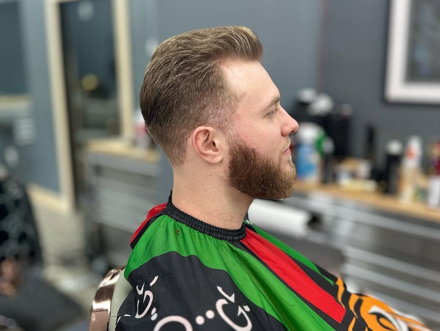 Side profile of a bearded man in a green and red barber cape seated in a barbershop.