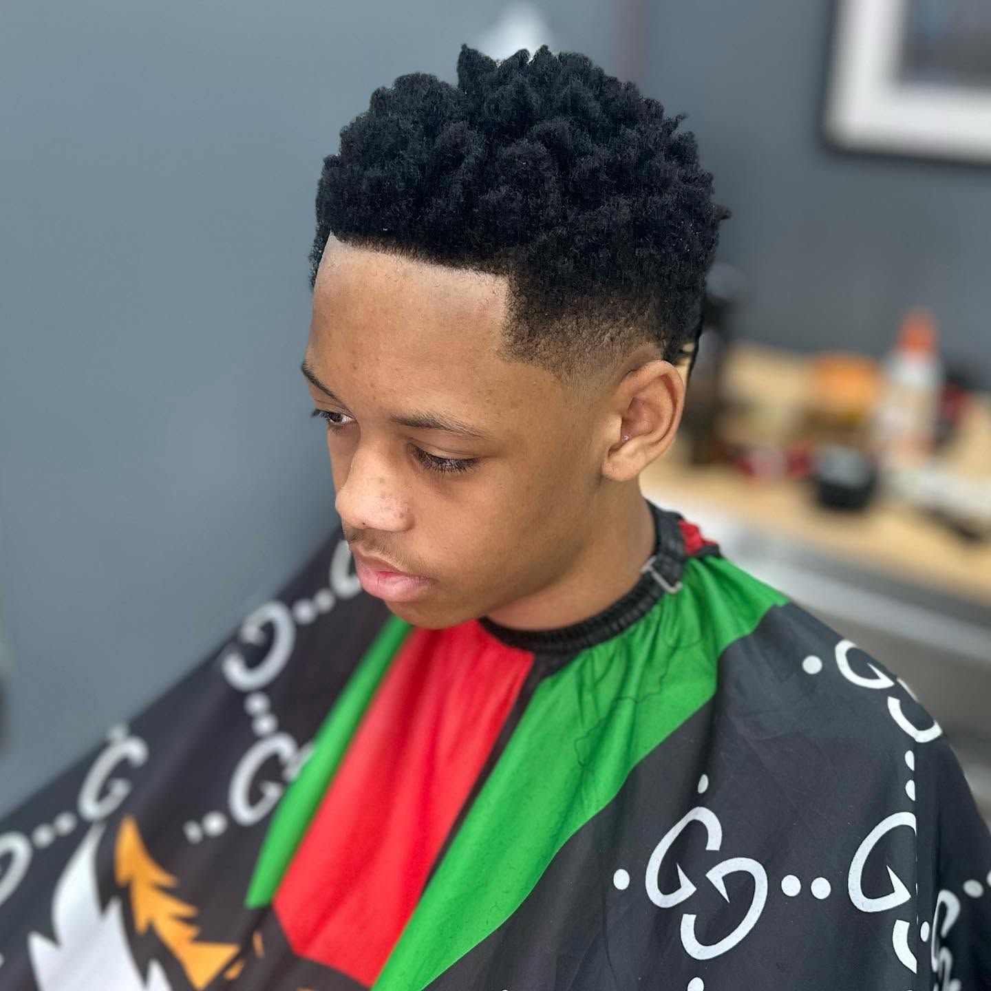 Young person getting a haircut in a barber chair, wearing a black cape with green and red trim.