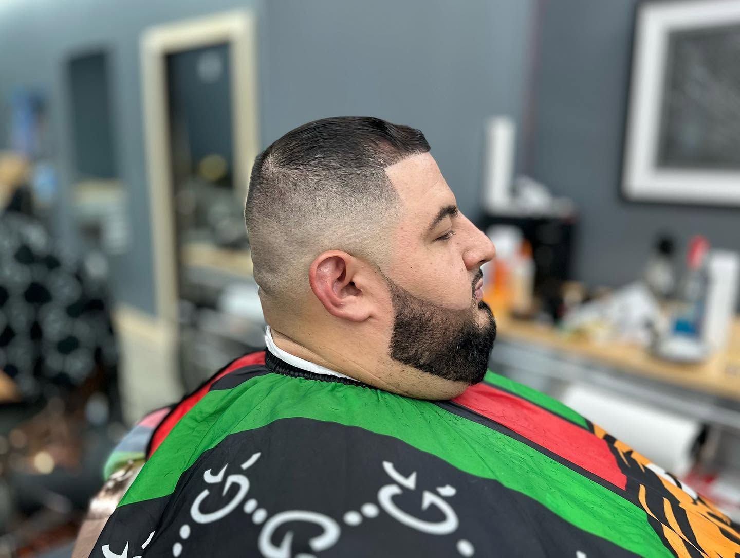 Man in a barber chair, side profile with a fresh fade haircut and a green striped cape in a salon.