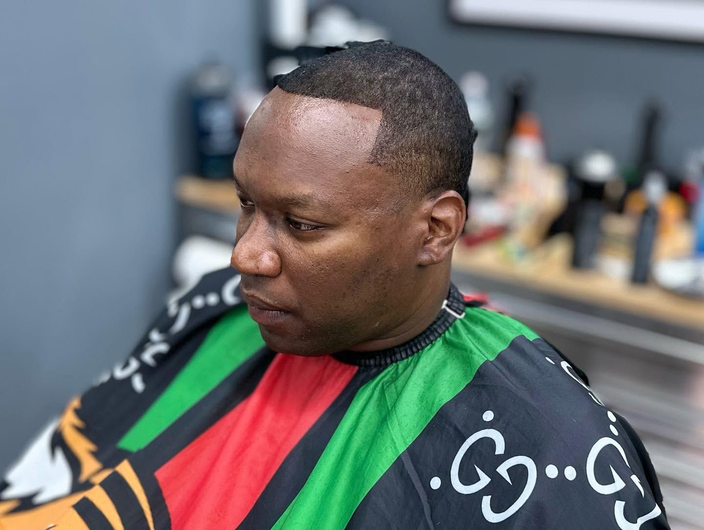Man seated in a barber chair wearing a colorful cape, looking down in a barbershop.