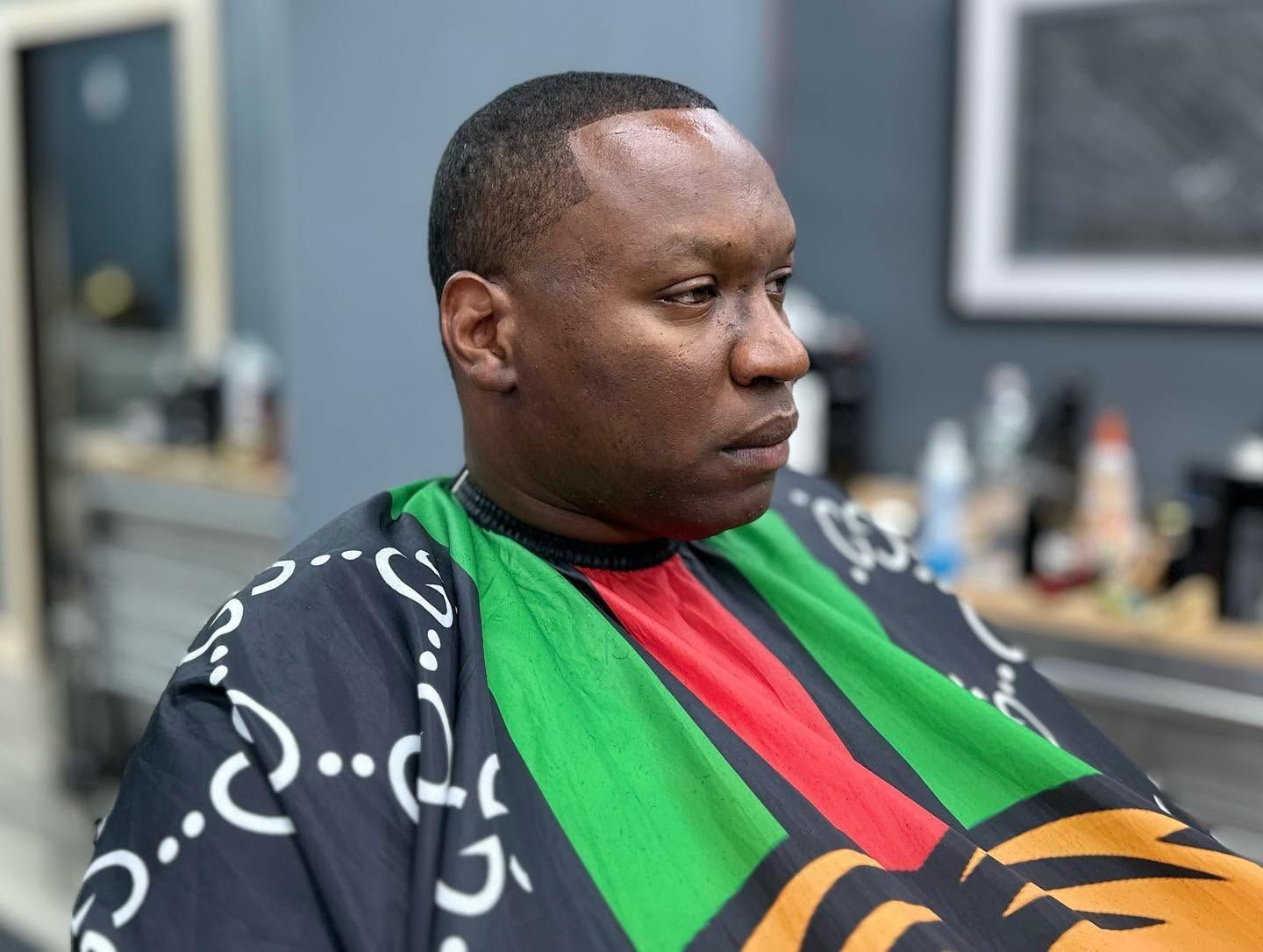 Person sitting in a salon chair, wearing a black patterned cape with green, red, and orange sections.