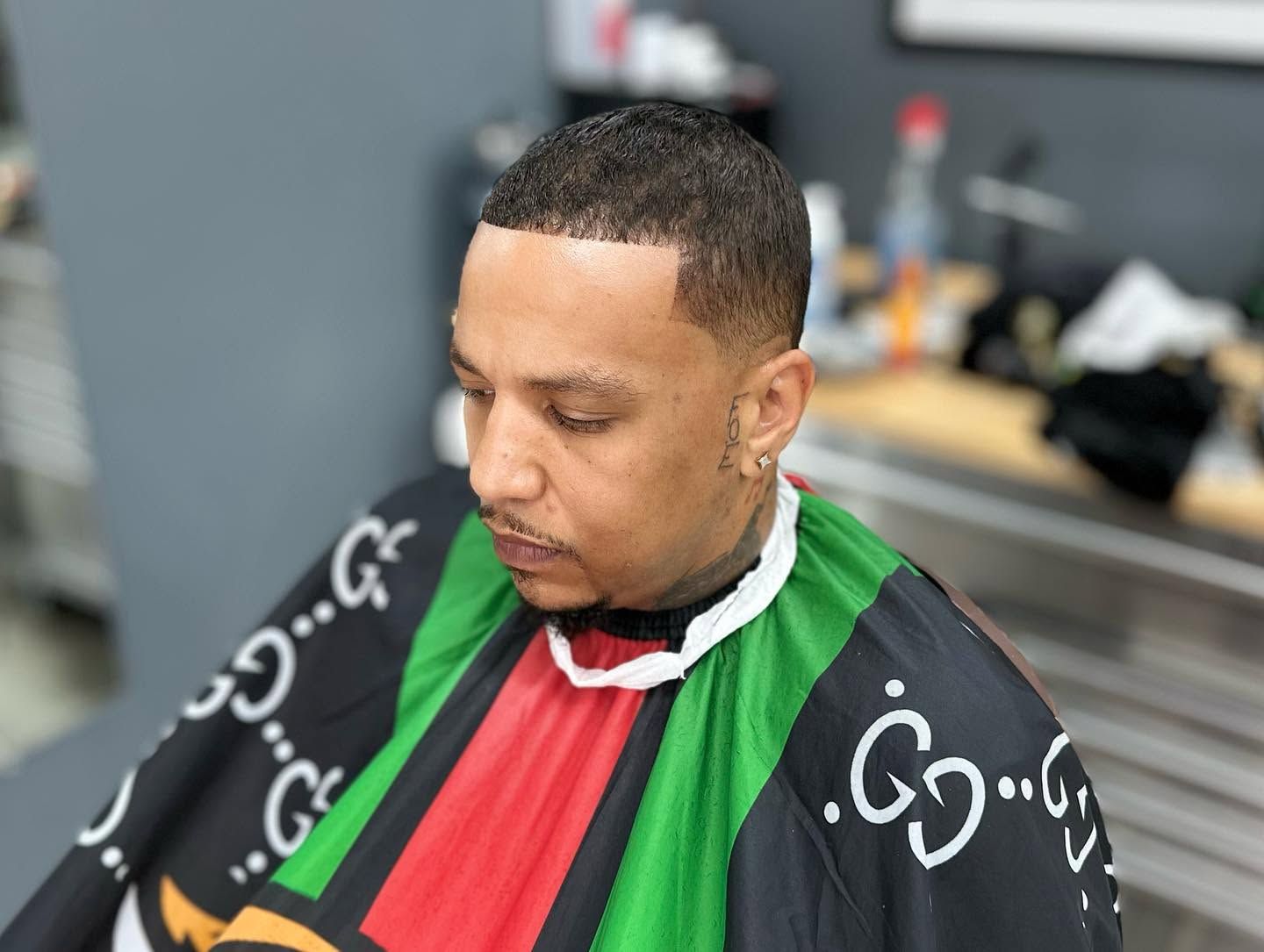 Man in a barber chair wearing a black, green, and red cape, looking down in a barbershop