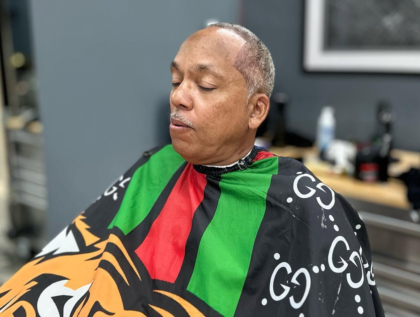Person seated in a barber chair wearing a black, green, and red cape with a tiger design, eyes closed.