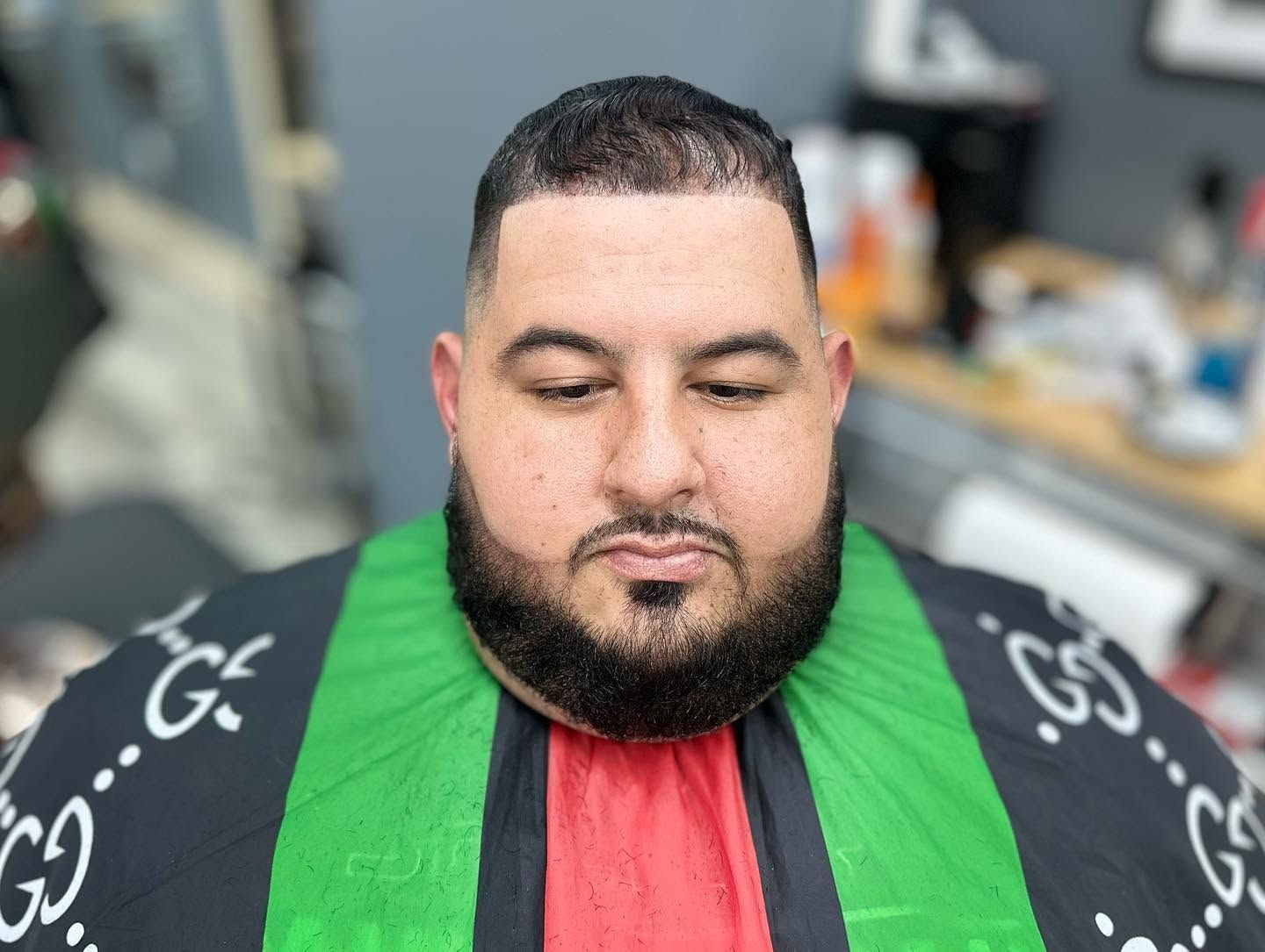 Man seated in a barber chair, wearing a black cape with green and red stripes, looking down with eyes closed.