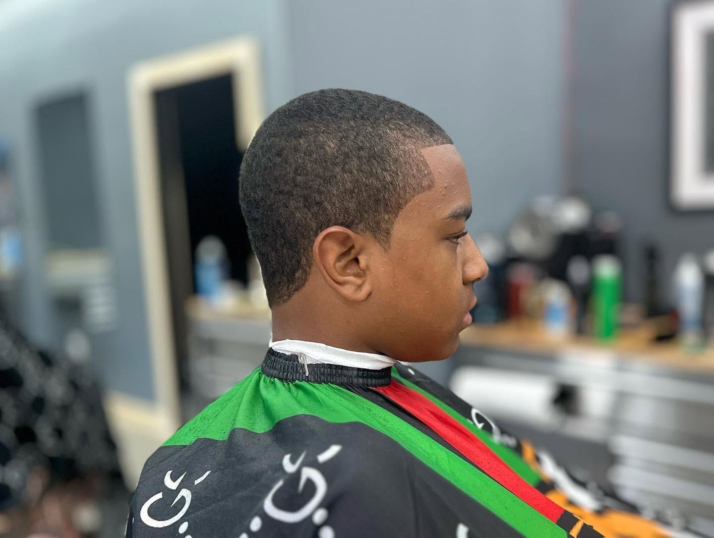 Person in a barbershop wearing a black cape with green and red trim, viewed in profile.
