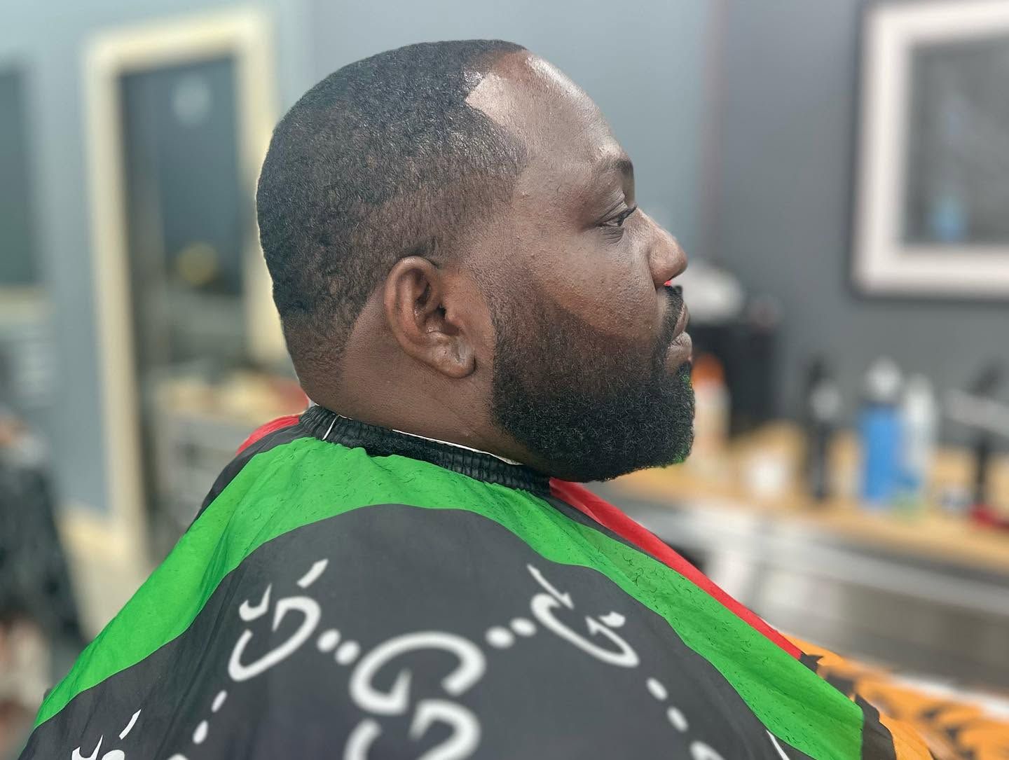 Man seated in a barber chair wearing a green and black barber cape in a salon