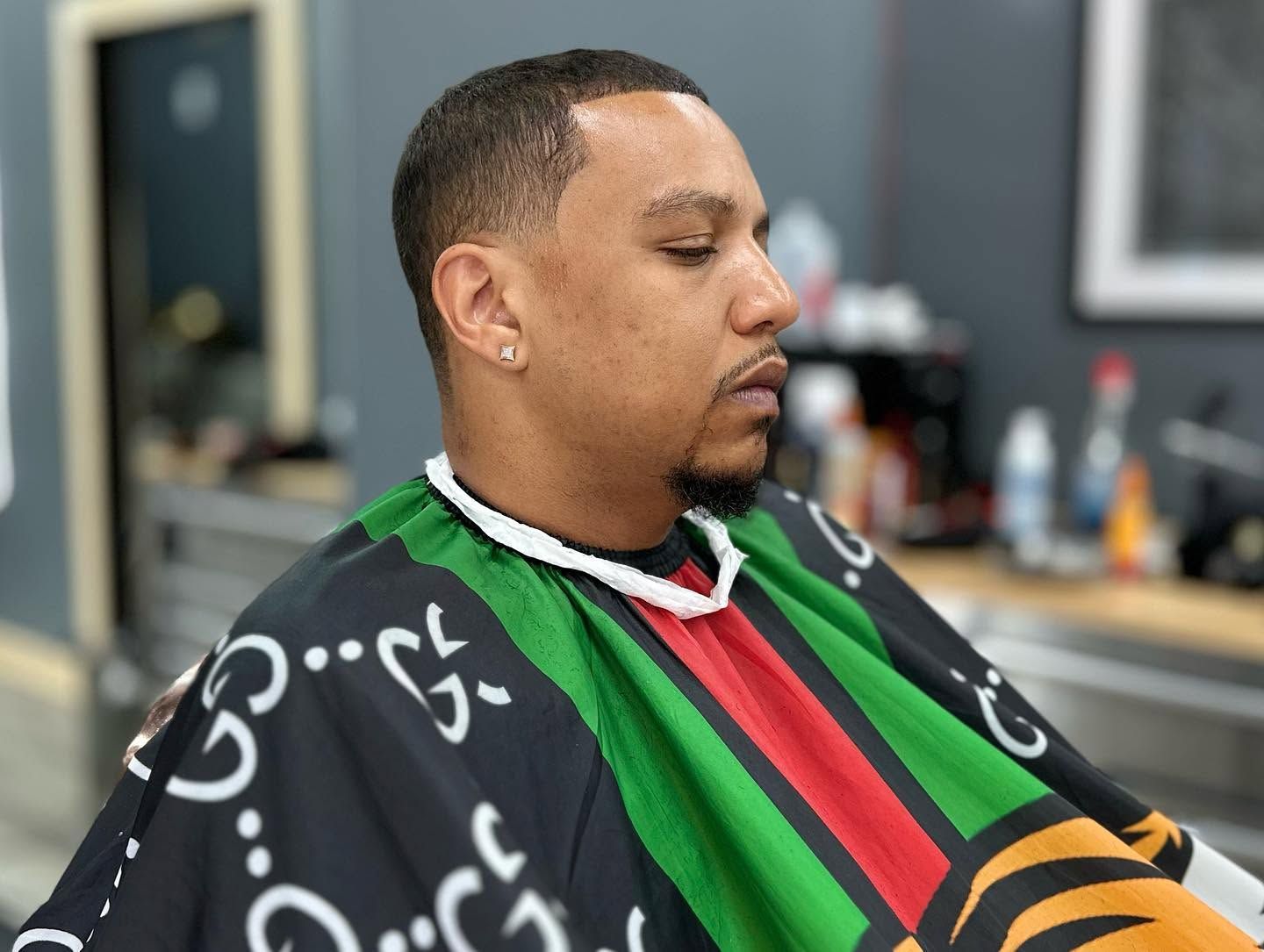 Man in a barber chair wearing a black, green, and red cape, getting a haircut in a barbershop.