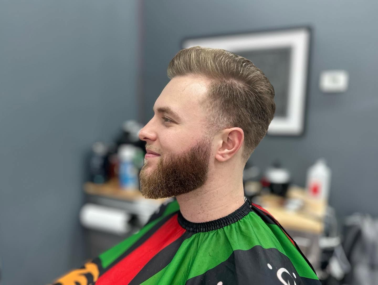 Man with a beard sitting in a barber chair, wearing a red and green cape in a salon