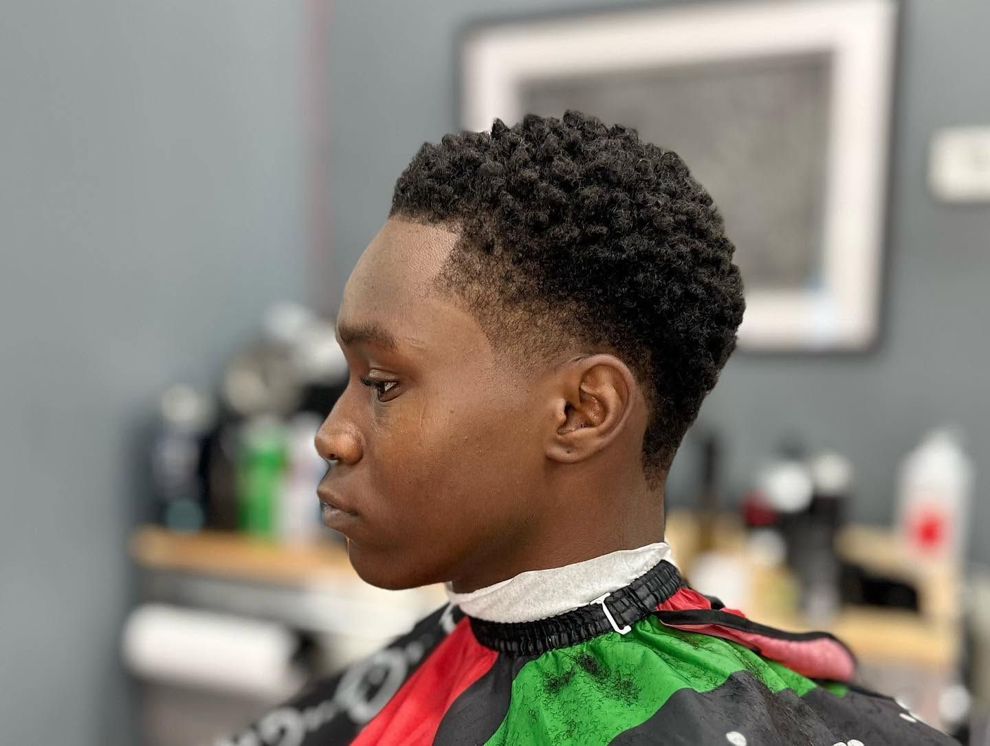 Profile of a person in a green and red barber cape, seated in a salon with a blurred mirror in the background