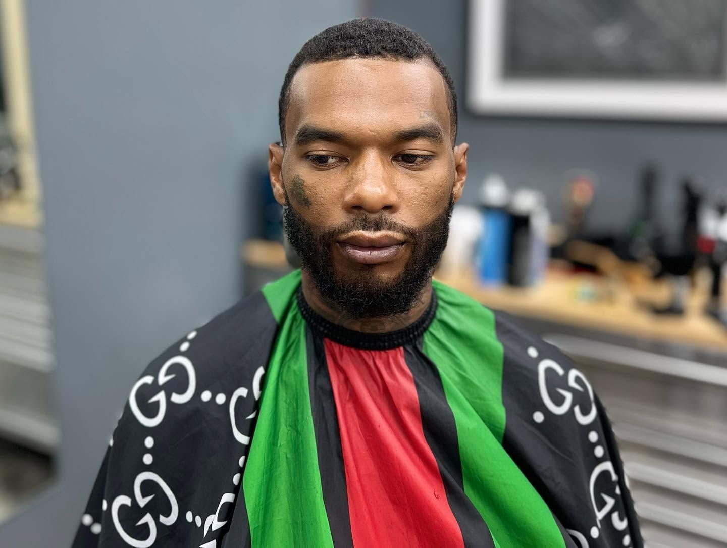 Man seated indoors wearing a black, green, and red striped barber cape, looking down thoughtfully