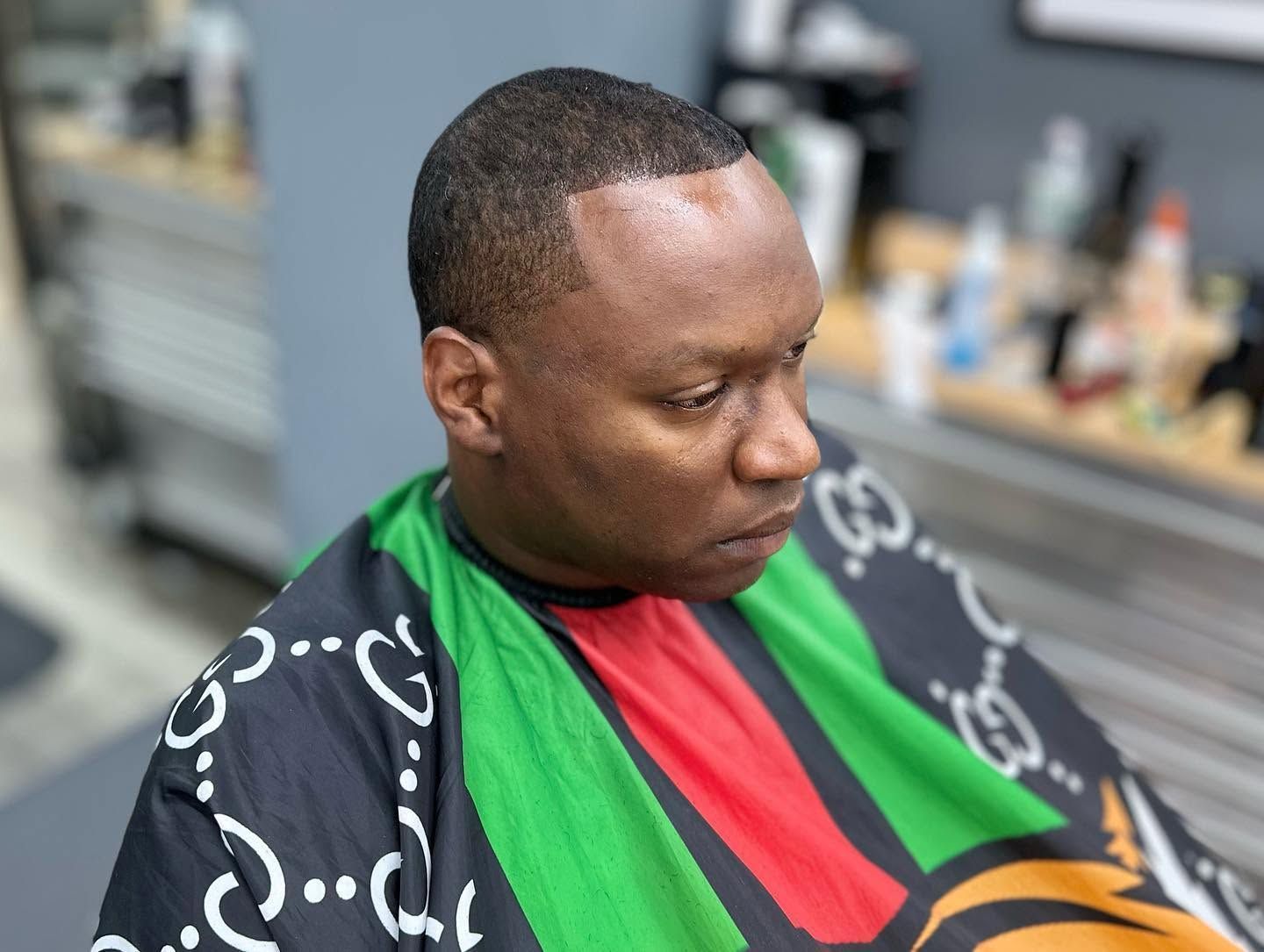 Man seated in a barber chair wearing a black, green, red, and yellow cape, looking down to the side.