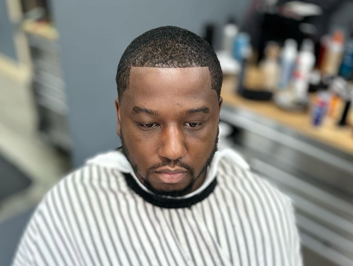 Man sitting in a barber chair wearing a striped cape, looking down in a barbershop.